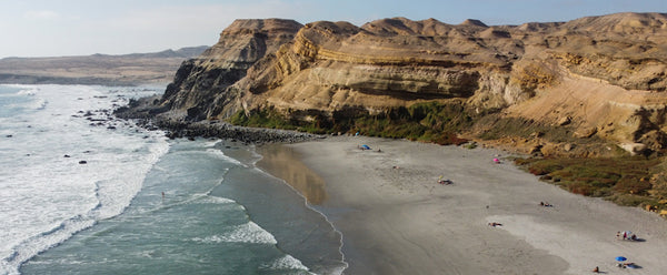 Playa entremedio de un acantilado, arena blanca y mar azul claro. Personas disfrutando del sol con quitasoles, sillas y toallas de playa.